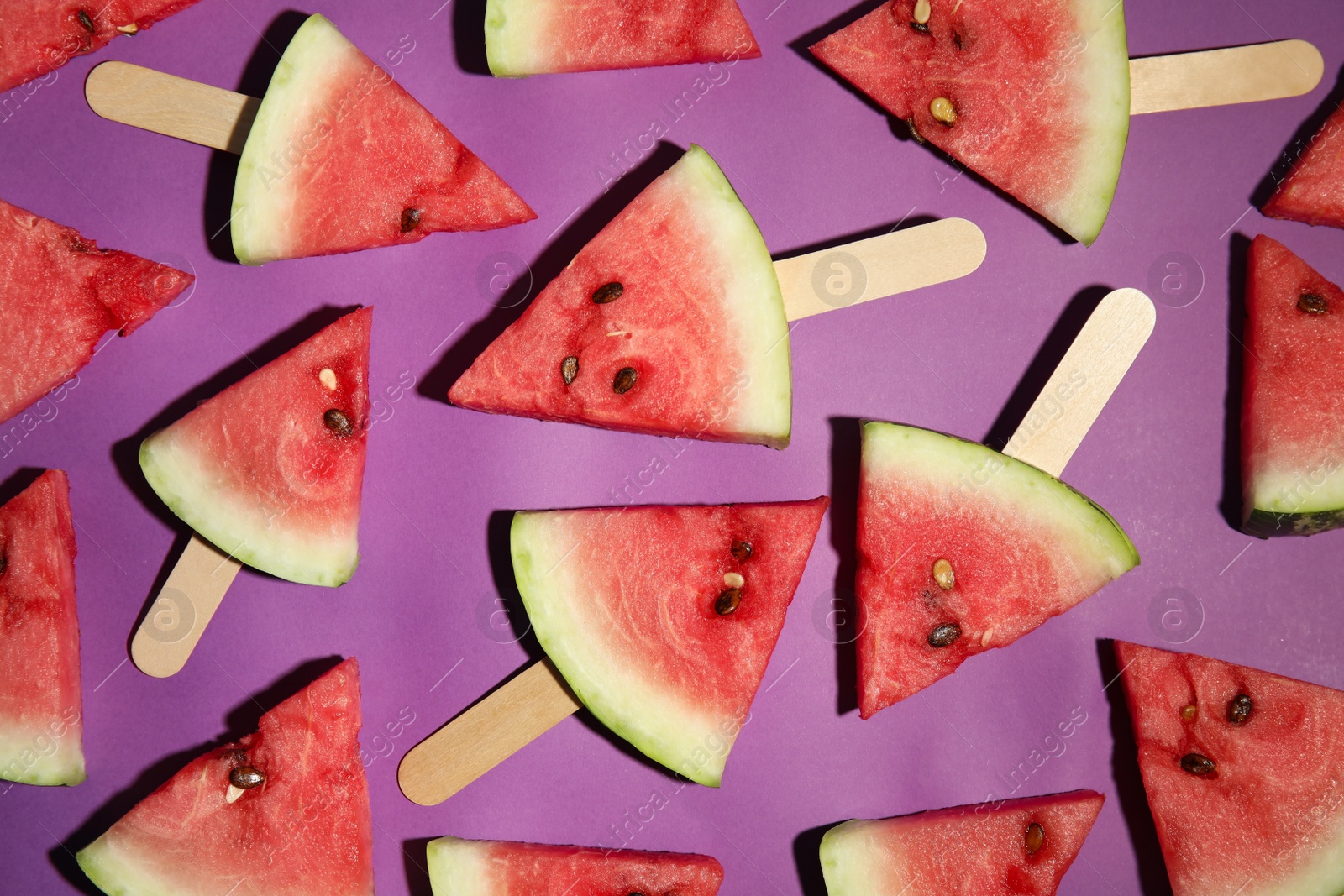 Photo of Watermelon slices with wooden sticks on purple background, flat lay