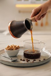 Woman pouring coffee from turkish pot into glass cup at white table, closeup