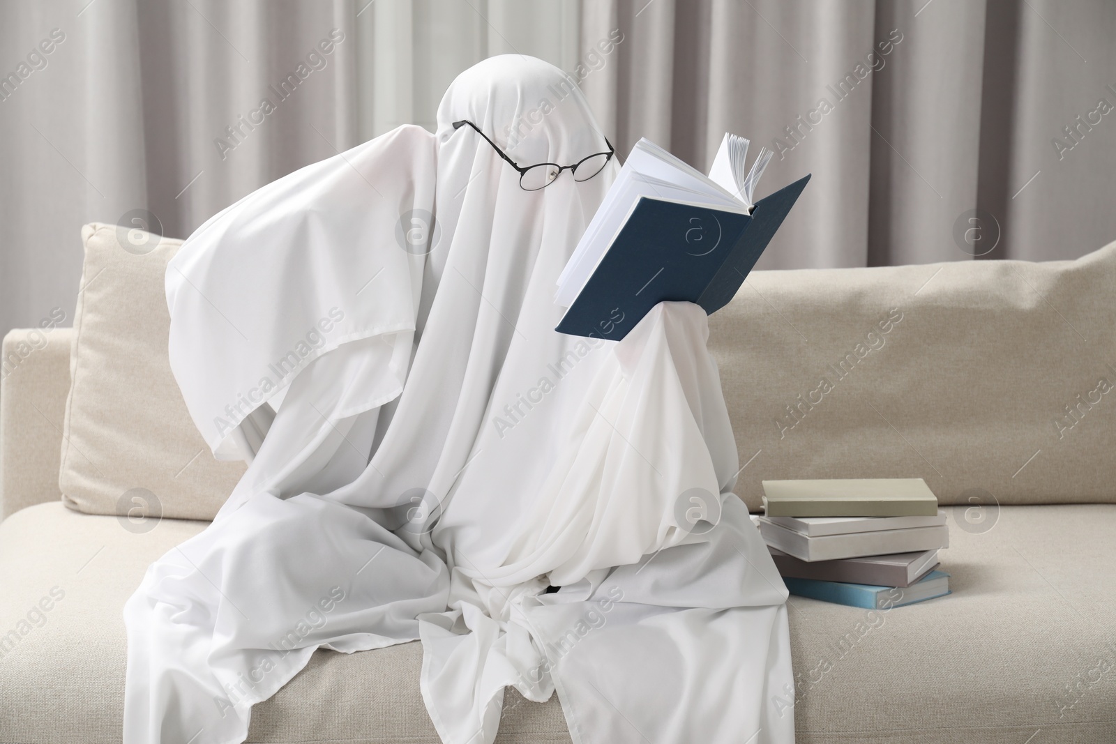 Photo of Creepy ghost. Person covered with white sheet reading book on sofa at home