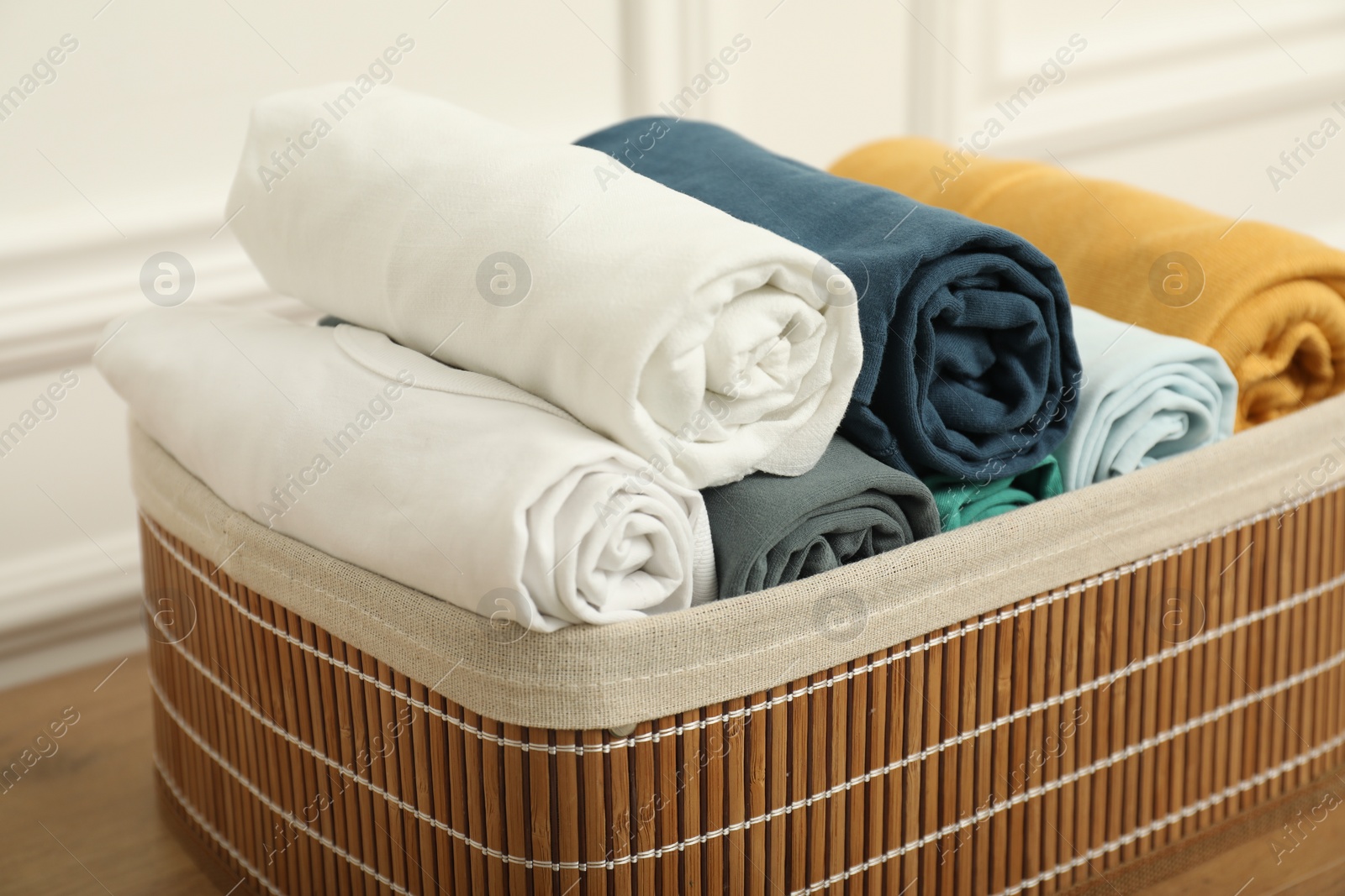 Photo of Basket with different rolled shirts on wooden table near white wall, closeup. Organizing clothes