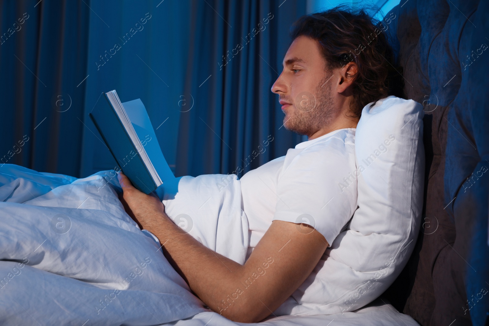 Photo of Handsome young man reading book in dark room at night. Bedtime