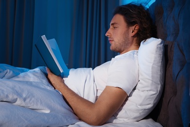 Photo of Handsome young man reading book in dark room at night. Bedtime