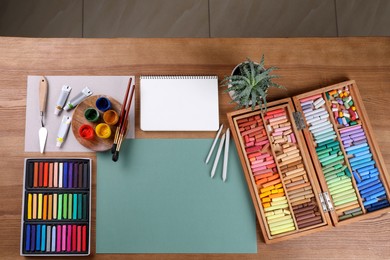 Blank sheet of paper, colorful chalk pastels and other drawing tools on wooden table, flat lay. Modern artist's workplace