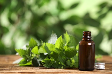 Glass bottle of nettle oil with fresh leaves on wooden table against blurred background, space for text