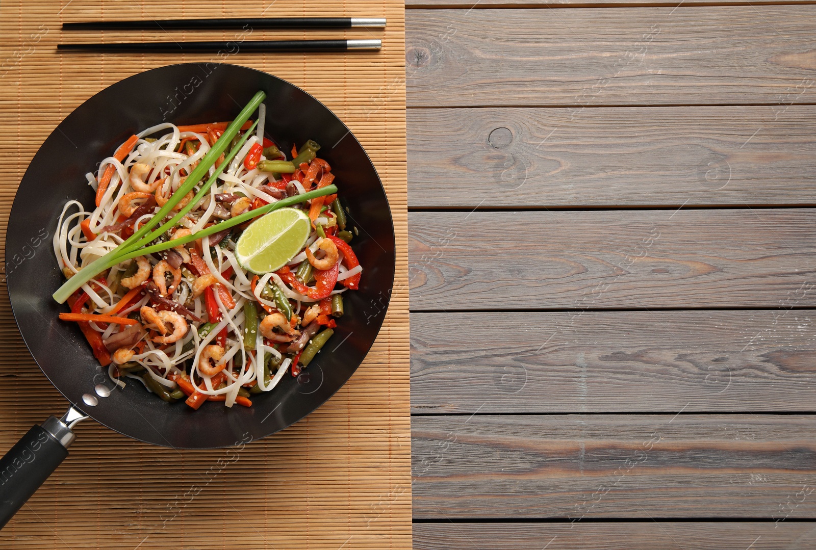 Photo of Shrimp stir fry with noodles and vegetables in wok on wooden table, top view. Space for text