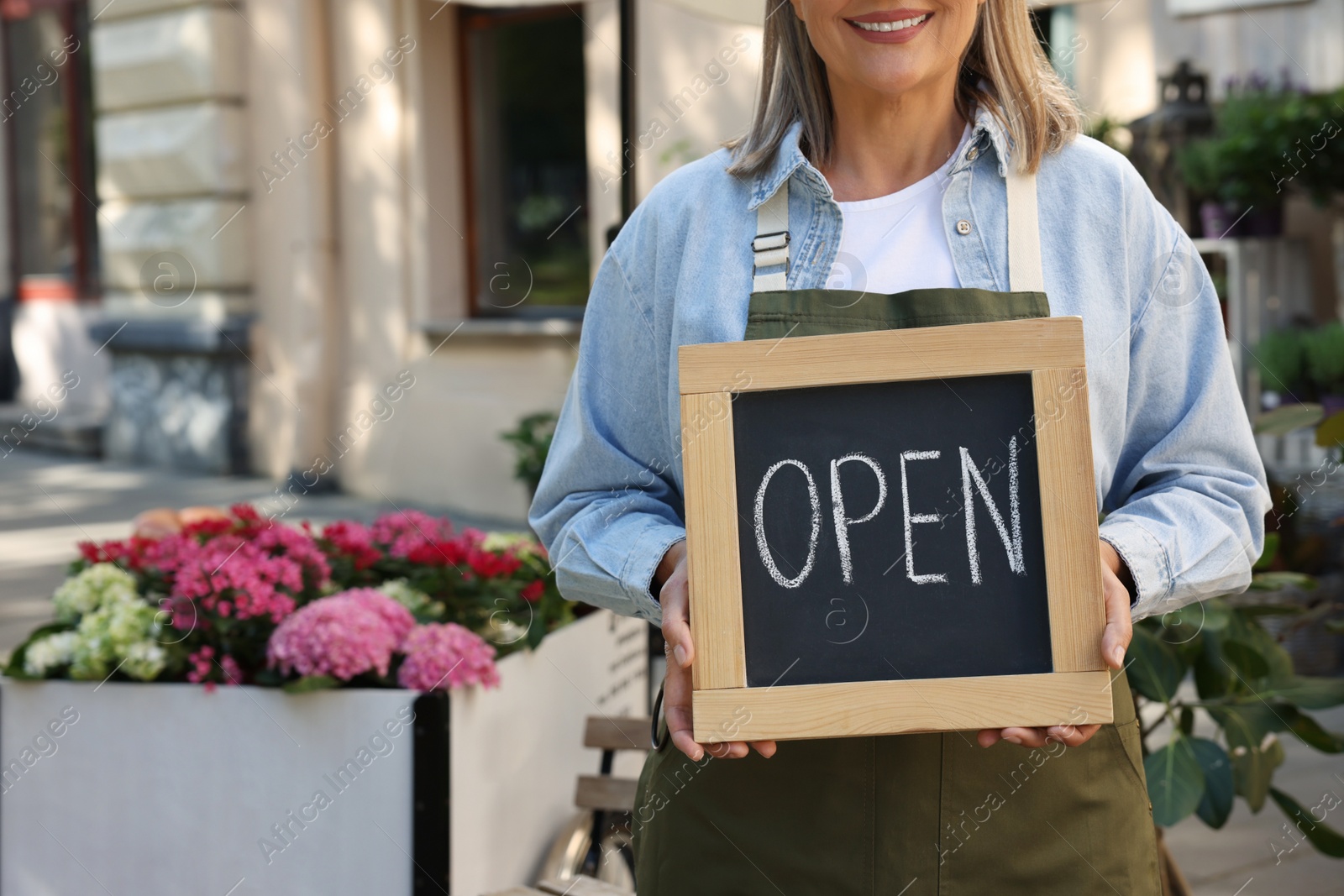 Photo of Business owner holding open sign near her flower shop outdoors, closeup. Space for text