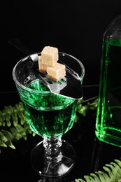 Photo of Absinthe in glass, brown sugar and spoon on mirror table, closeup. Alcoholic drink