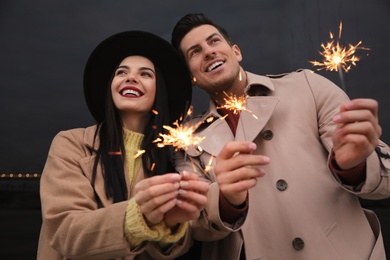 Couple in warm clothes holding burning sparklers near building