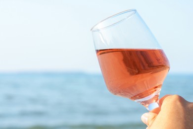 Woman holding glass of tasty rose wine near sea, closeup. Space for text