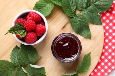 Photo of Jar of delicious raspberry jam, fresh berries and green leaves on table, flat lay