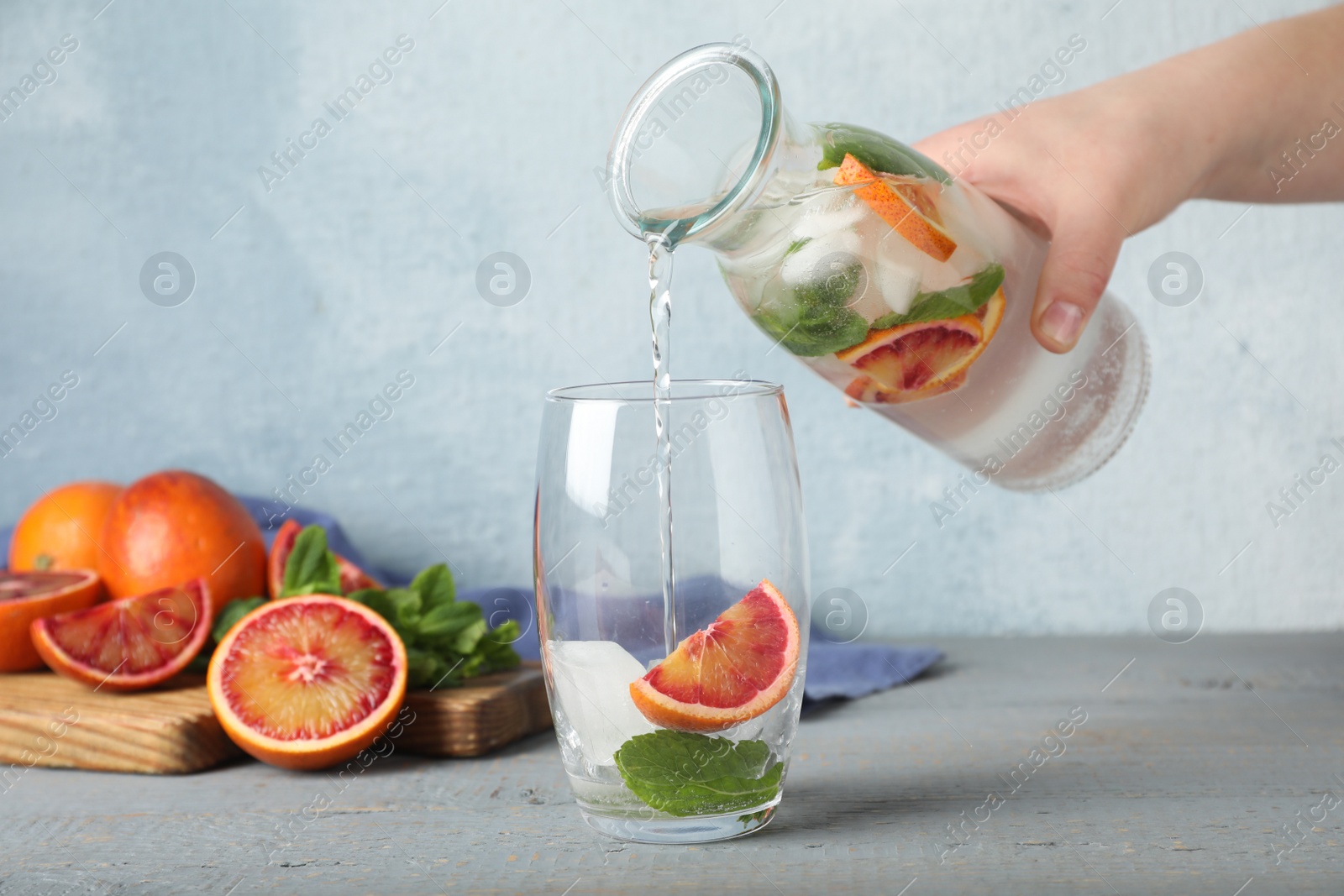 Photo of Woman pouring refreshing drink with sicilian orange from bottle into glass at grey wooden table, closeup