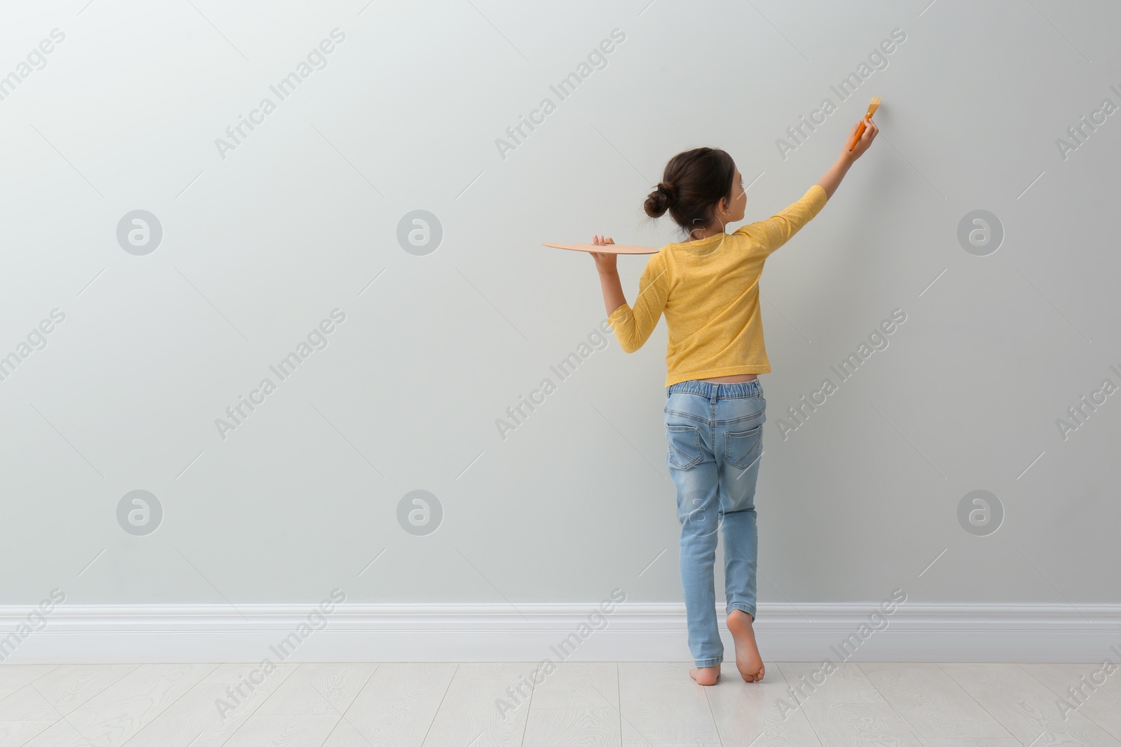 Photo of Little girl painting on light grey wall indoors, back view. Space for text