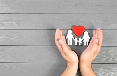 Photo of Young man protecting family figure and red heart with his hands on grey wooden background, top view. Space for text