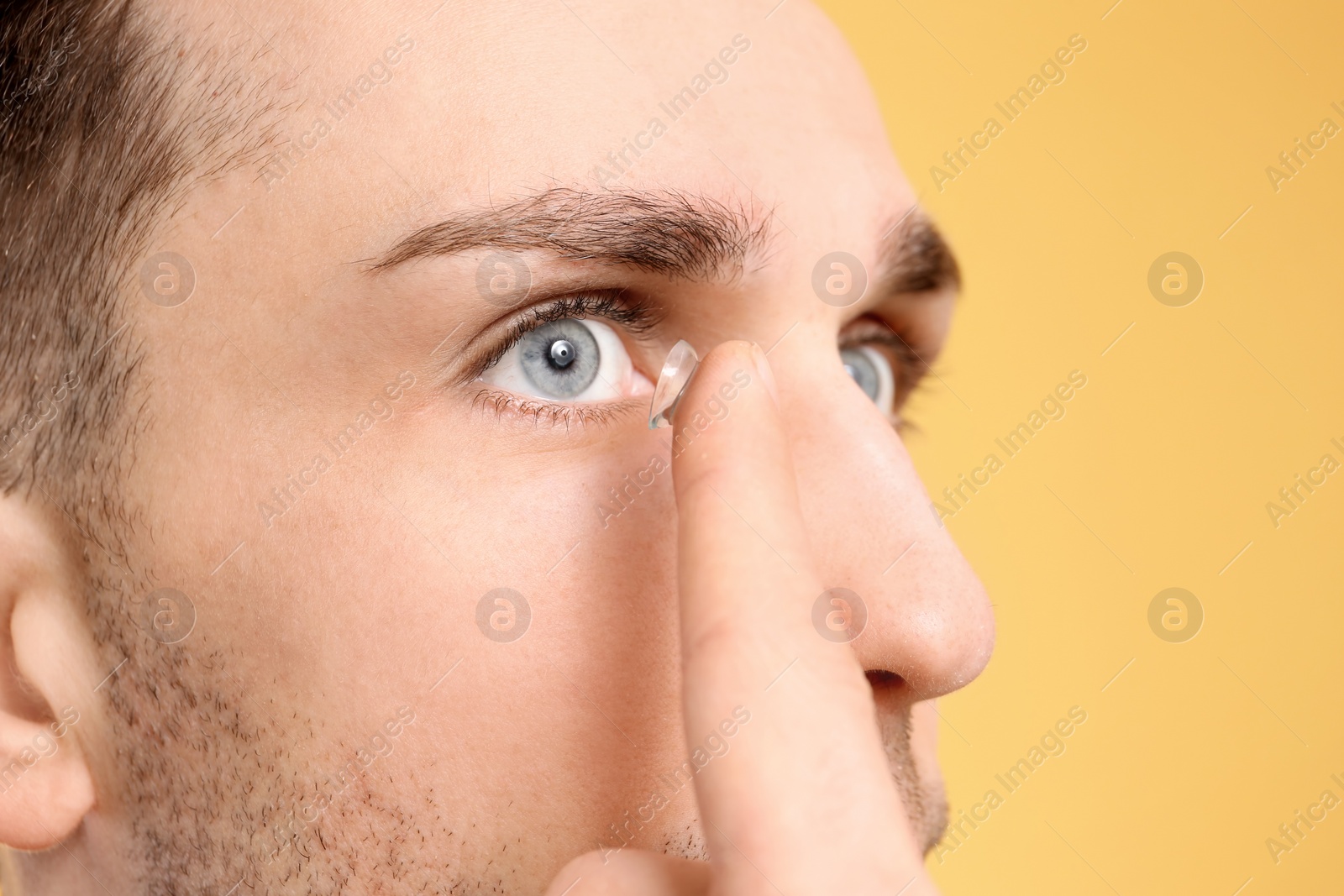 Photo of Young man putting contact lens in his eye on color background