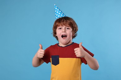 Photo of Birthday celebration. Cute little boy in party hat showing thumbs up on light blue background