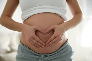 Pregnant woman making heart with her hands near belly indoors, closeup