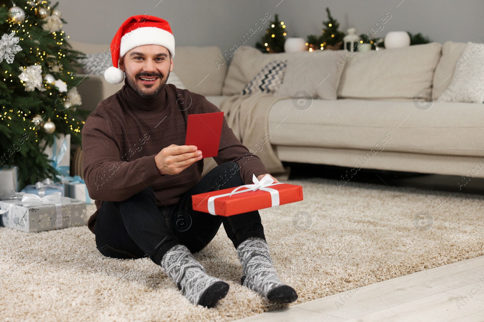 Photo of Happy man in Santa hat with Christmas gift and greeting card at home