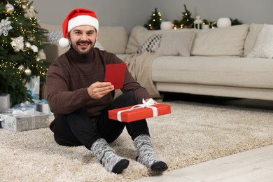 Happy man in Santa hat with Christmas gift and greeting card at home