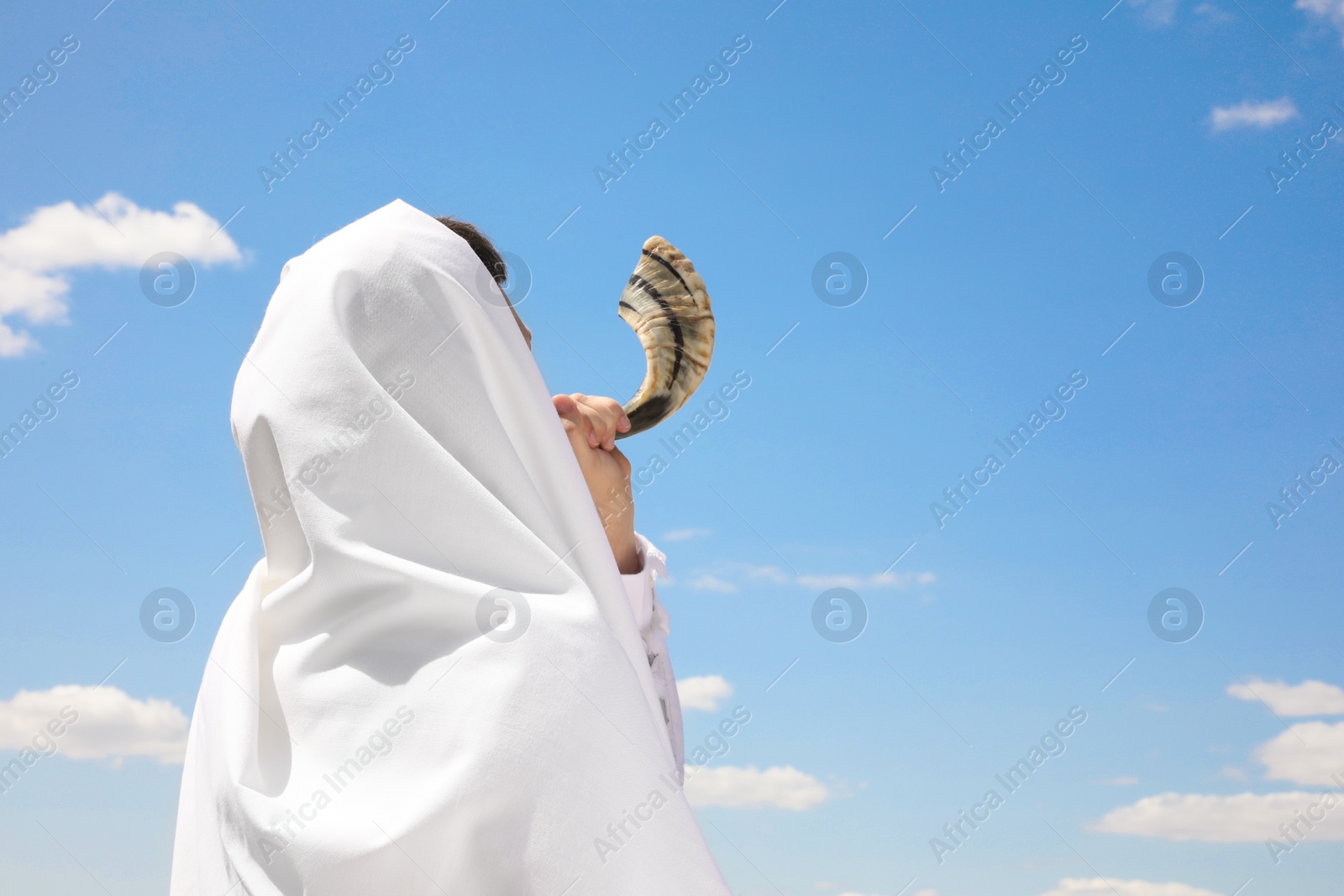 Photo of Jewish man in tallit blowing shofar outdoors. Rosh Hashanah celebration