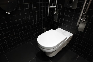 Photo of Clean ceramic toilet bowl and handrails on tiled wall indoors