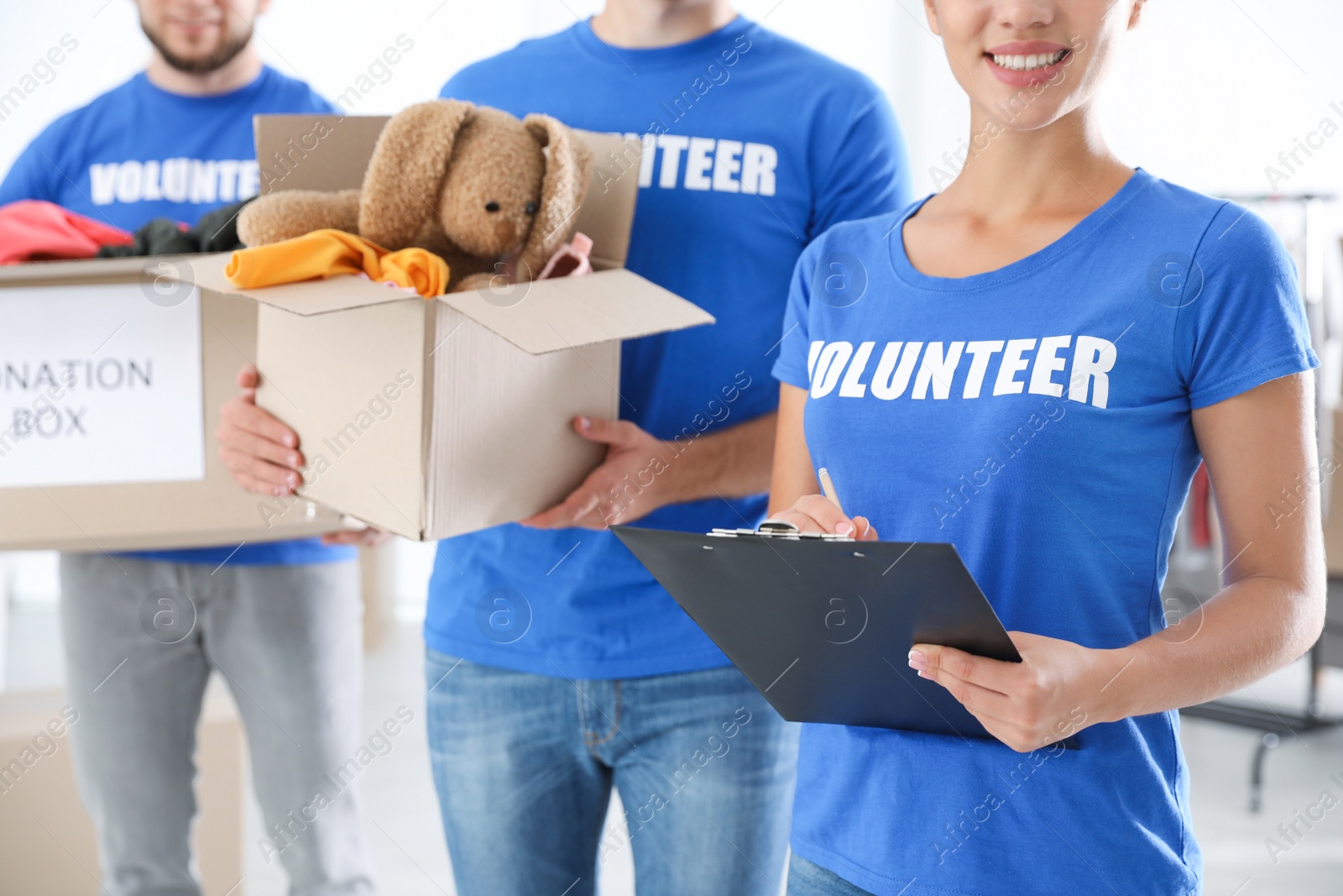 Photo of Young female volunteer with clipboard and her team holding boxes full of donations indoors