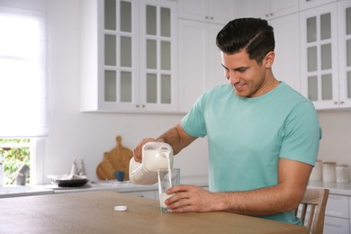 Photo of Man pouring milk from gallon bottle into glass at wooden table in kitchen