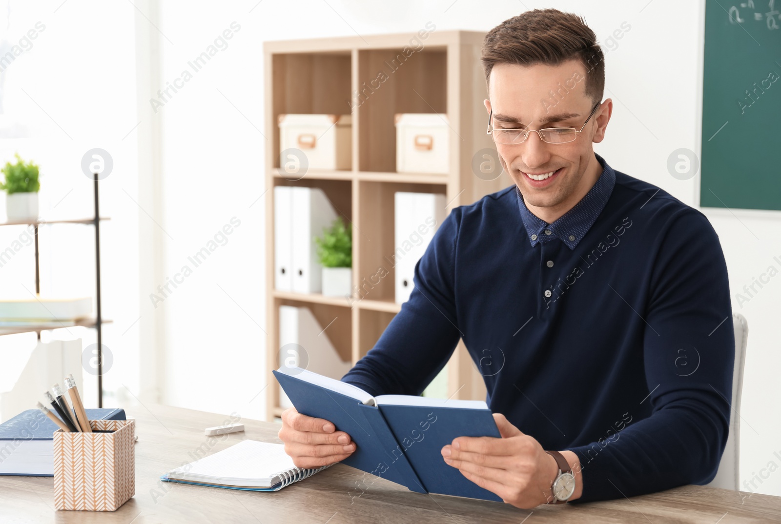 Photo of Young male teacher with book sitting at table in classroom