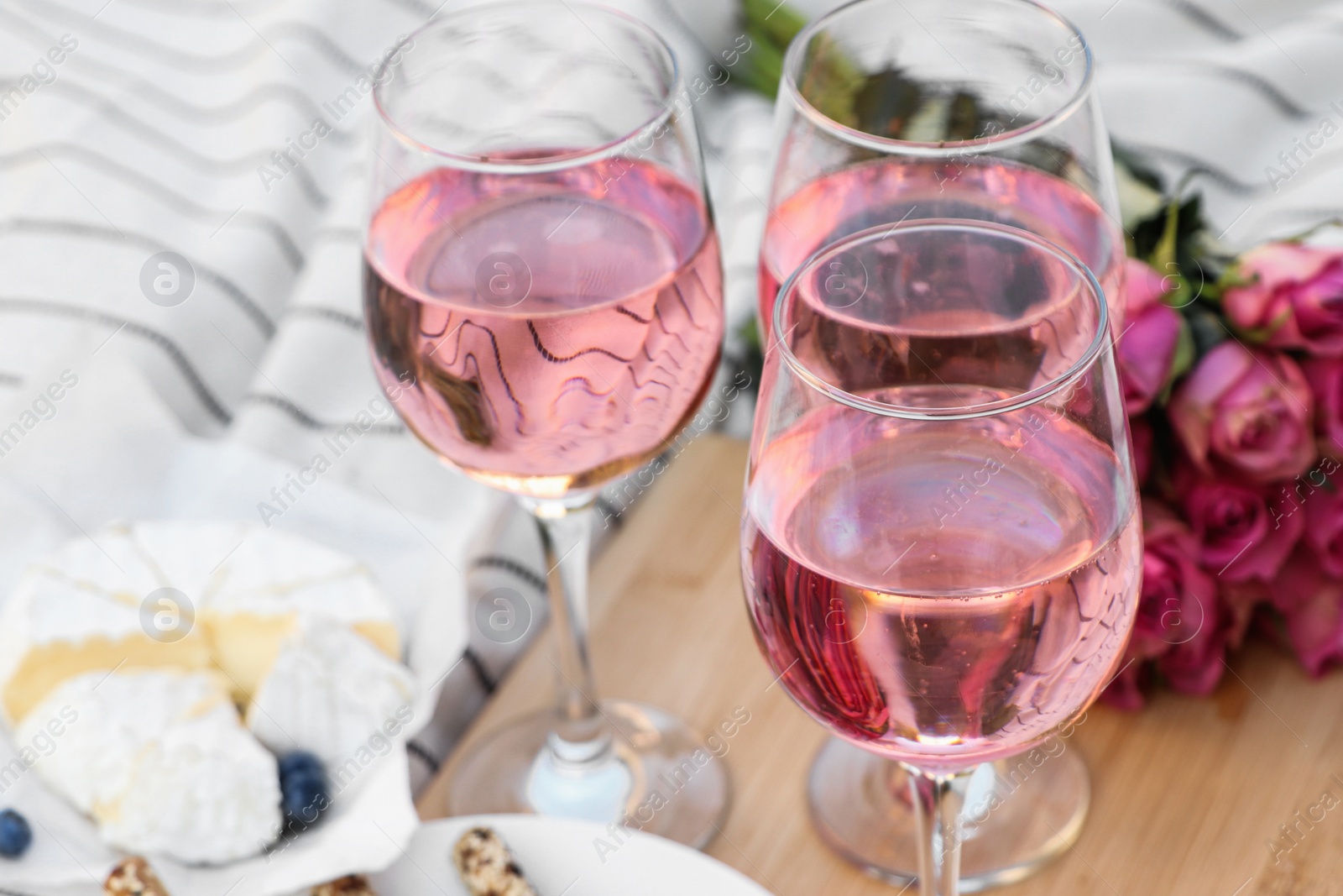 Photo of Glasses of delicious rose wine, flowers and food on white picnic blanket, closeup