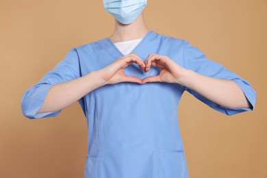 Photo of Nurse wearing protective mask and medical uniform making heart with hands on light brown background, closeup