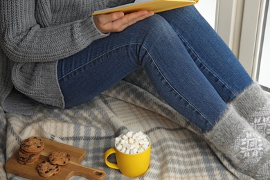 Photo of Woman with cup of hot cocoa reading book on window sill, closeup. Winter drink