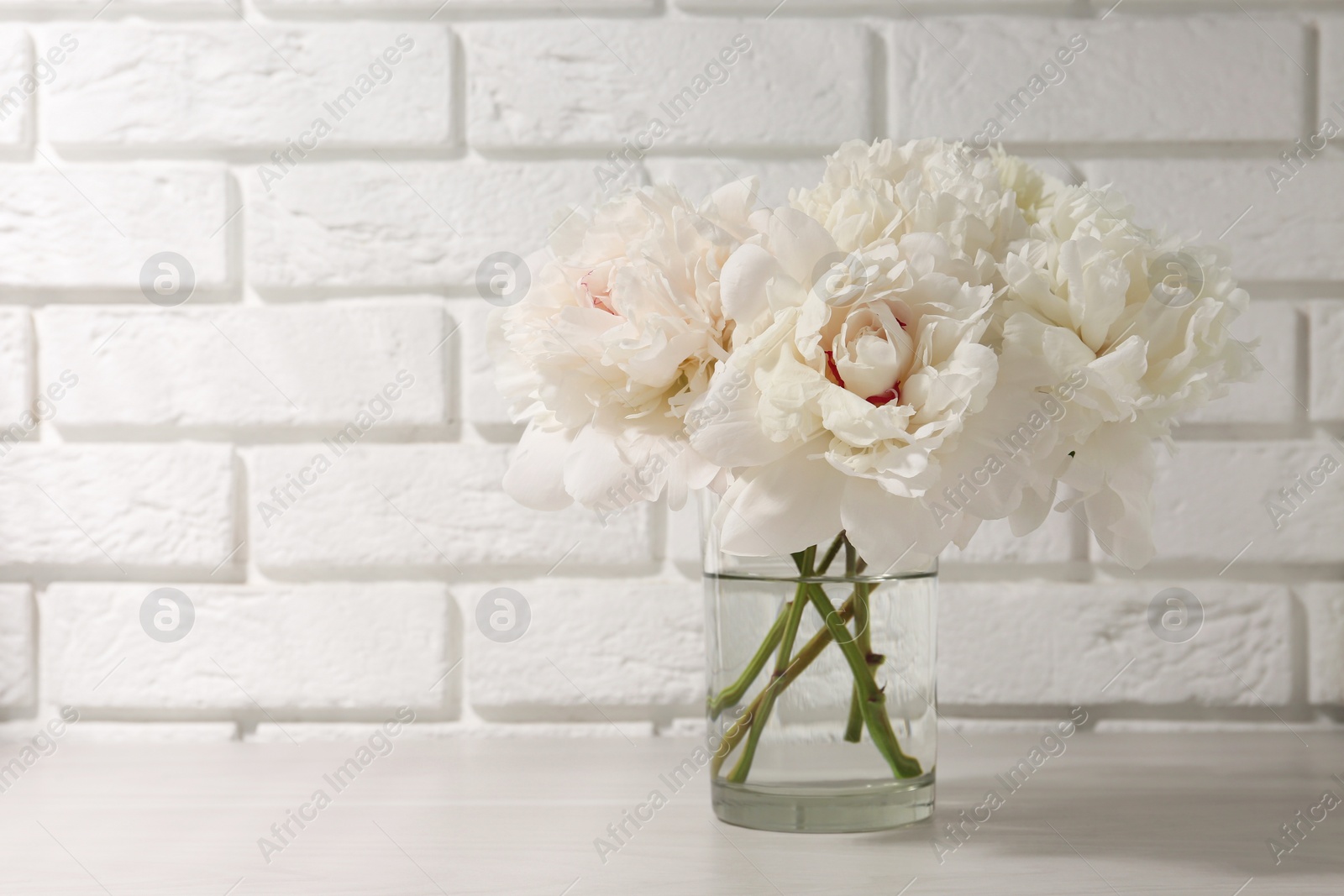 Photo of Beautiful peonies in glass vase on white table near brick wall. Space for text