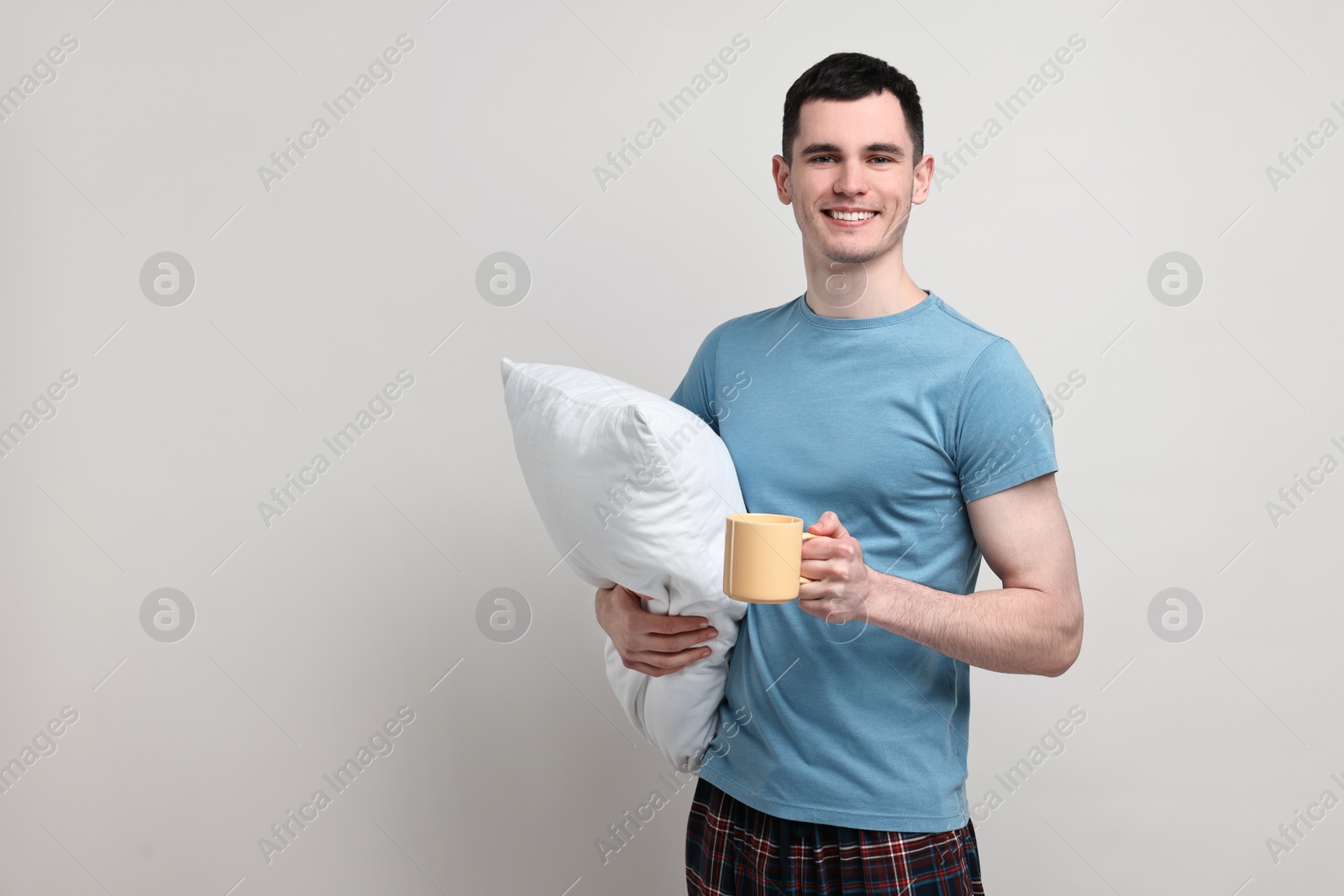 Photo of Happy man in pyjama holding pillow and cup of drink on light grey background, space for text