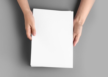 Woman holding blank paper sheets for brochure at grey table, top view. Mock up