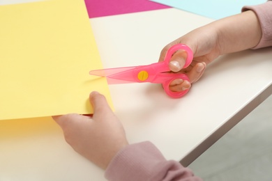 Photo of Child cutting color paper with plastic scissors at table, closeup