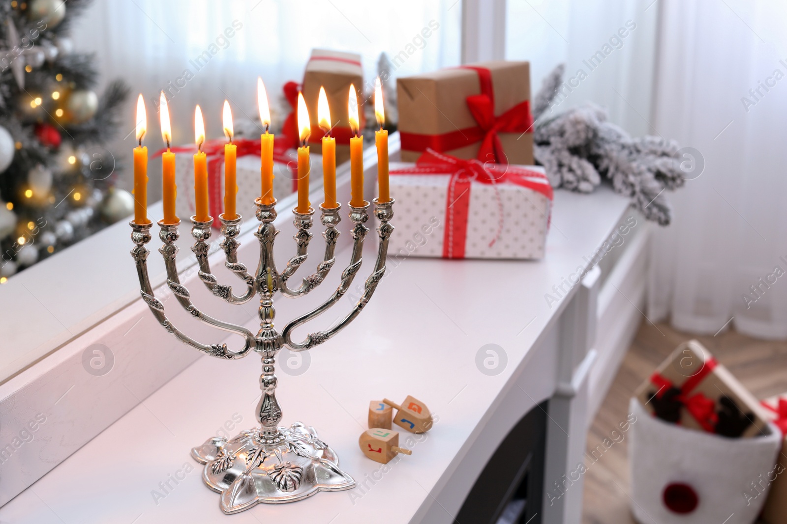 Photo of Silver menorah near dreidels with symbols He, Pe, Nun, Gimel on white mantelpiece in room decorated for Christmas