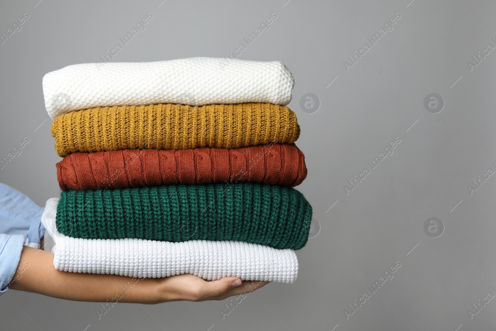 Photo of Woman holding stack of warm clothes on grey background, closeup. Autumn season