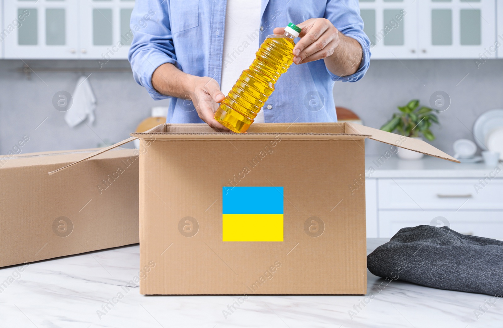 Photo of Humanitarian aid for Ukraine. Man putting bottle of oil in donation box at white table indoors, closeup