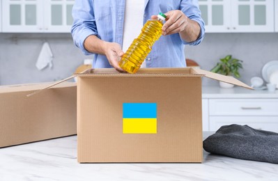 Photo of Humanitarian aid for Ukraine. Man putting bottle of oil in donation box at white table indoors, closeup