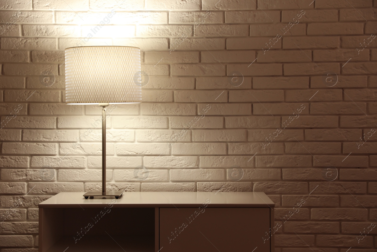 Photo of Stylish glowing night lamp on table near white brick wall. Space for text