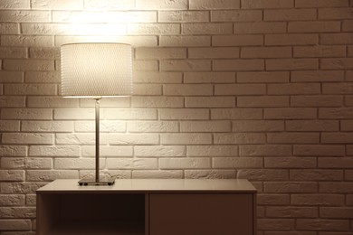 Stylish glowing night lamp on table near white brick wall. Space for text