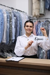 Photo of Dry-cleaning service. Happy worker holding Open sign at counter indoors