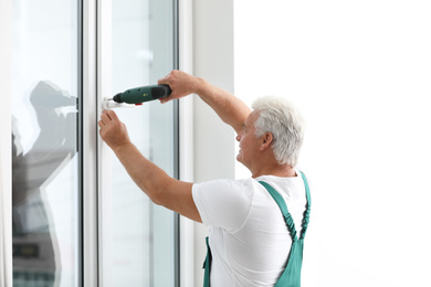 Mature construction worker repairing plastic window with electric screwdriver indoors