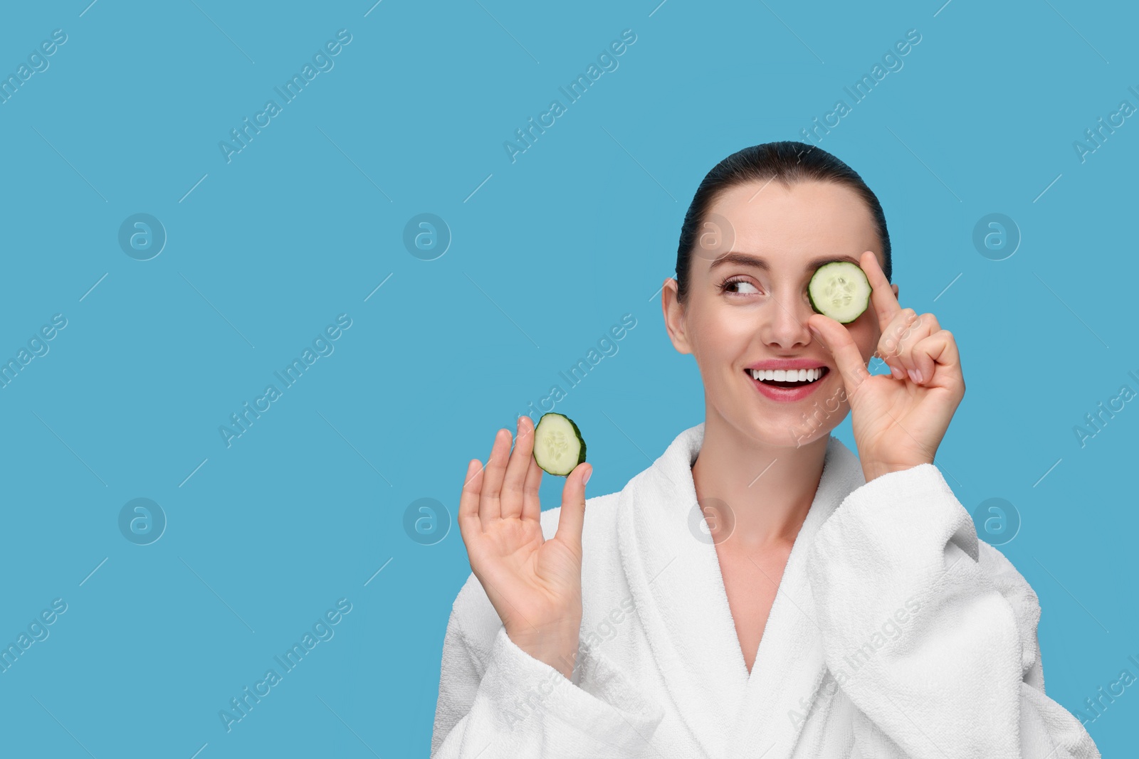 Photo of Beautiful woman covering eye with piece of cucumber on light blue background, space for text