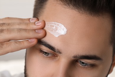 Man with dry skin applying cream onto his forehead on light background, closeup