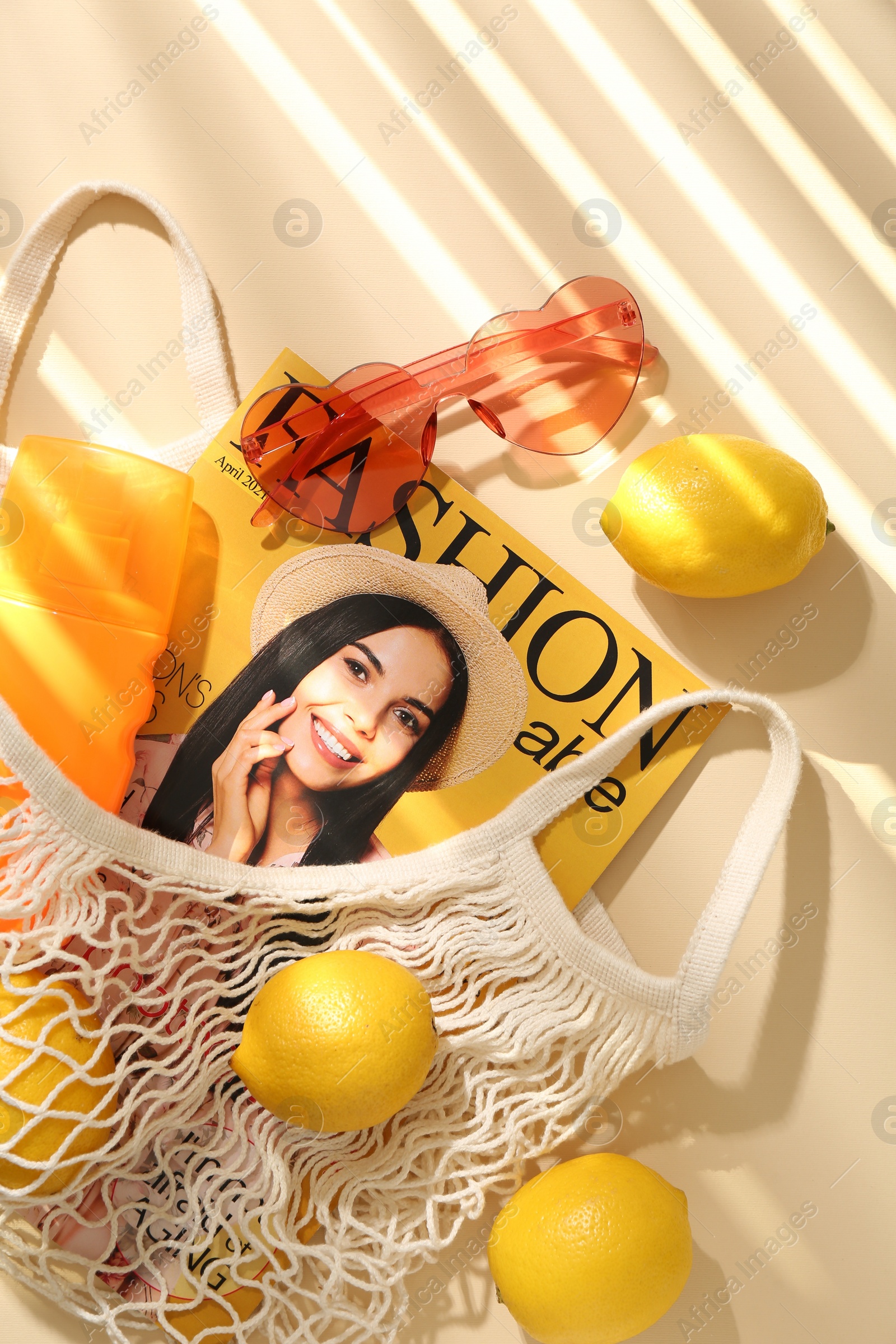 Photo of String bag with fresh lemons, fashion magazine and beach accessories on beige background, flat lay