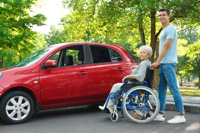 Young man with disabled senior woman in wheelchair near car outdoors