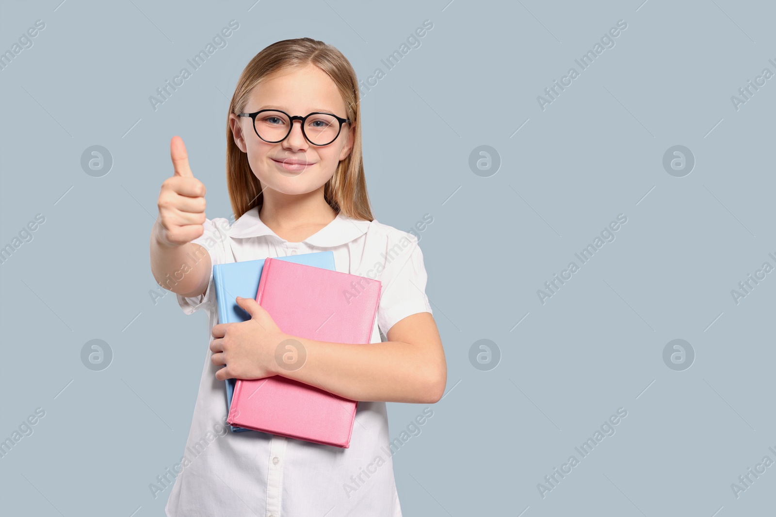Photo of Cute girl in glasses with books showing thumb up on light grey background. Space for text