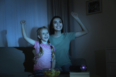 Emotional young woman and her daughter watching TV using video projector at home