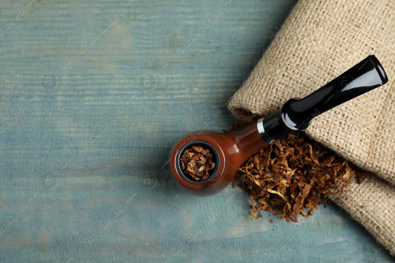 Photo of Smoking pipe, dry tobacco and sackcloth on blue wooden table, top view. Space for text