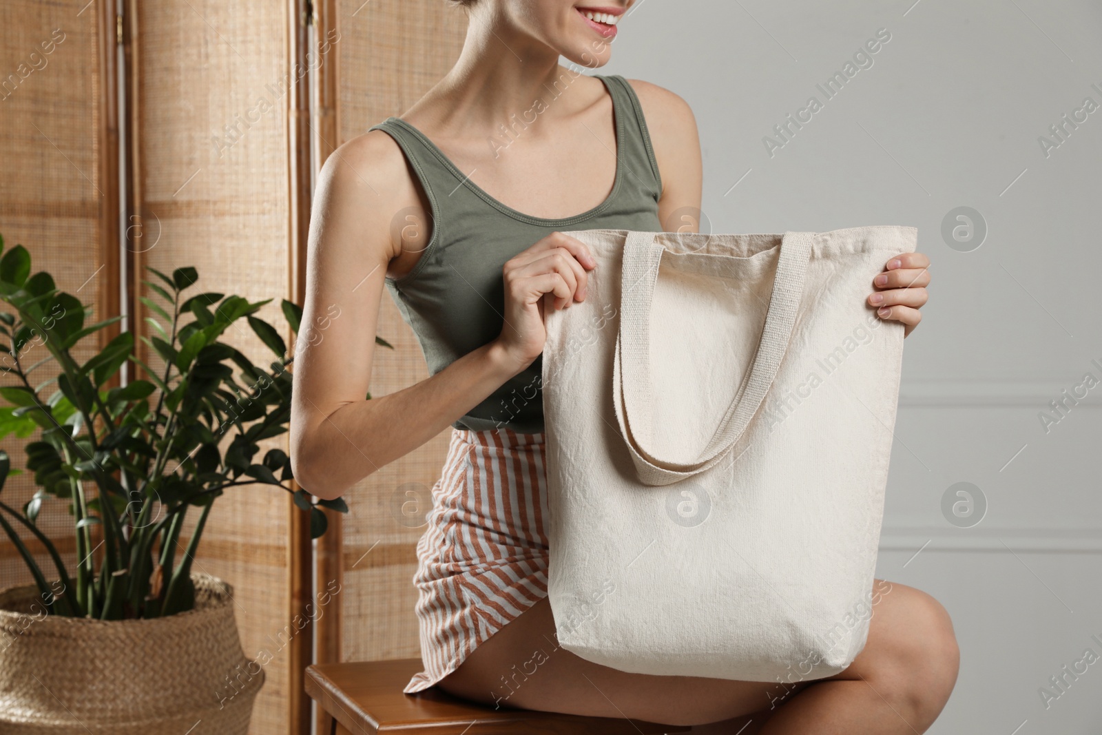 Photo of Happy young woman with blank eco friendly bag sitting on wooden stool indoors, closeup